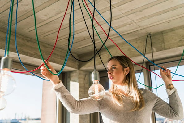 Junge Frau schaut auf Kabel an Decke vor Glühbirne symbolisiert Energiesparen im Büro