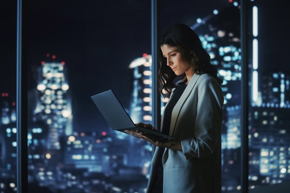 Frau arbeitet am Laptop mit Firewall vor dunkler Skyline hinter Fenster