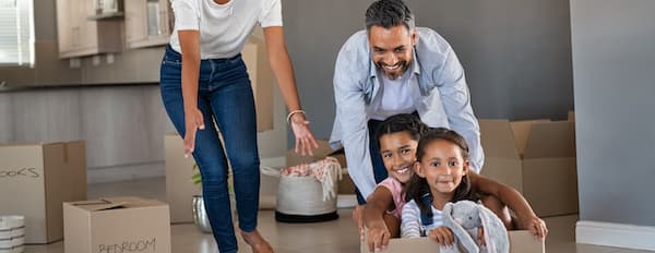 Smiling adult man pushing two smiling children in moving box as a woman reaches out her hands toward them. The room is filled with moving boxes.