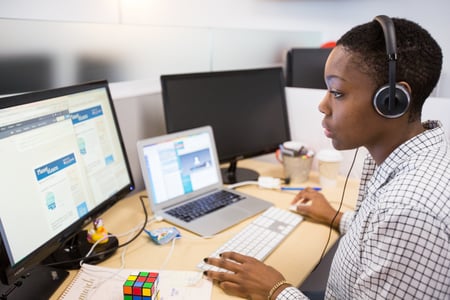 A picture of a woman wearing a headset, sitting at a desk in front of a desktop and laptop screen, looking through webpages and documents. 